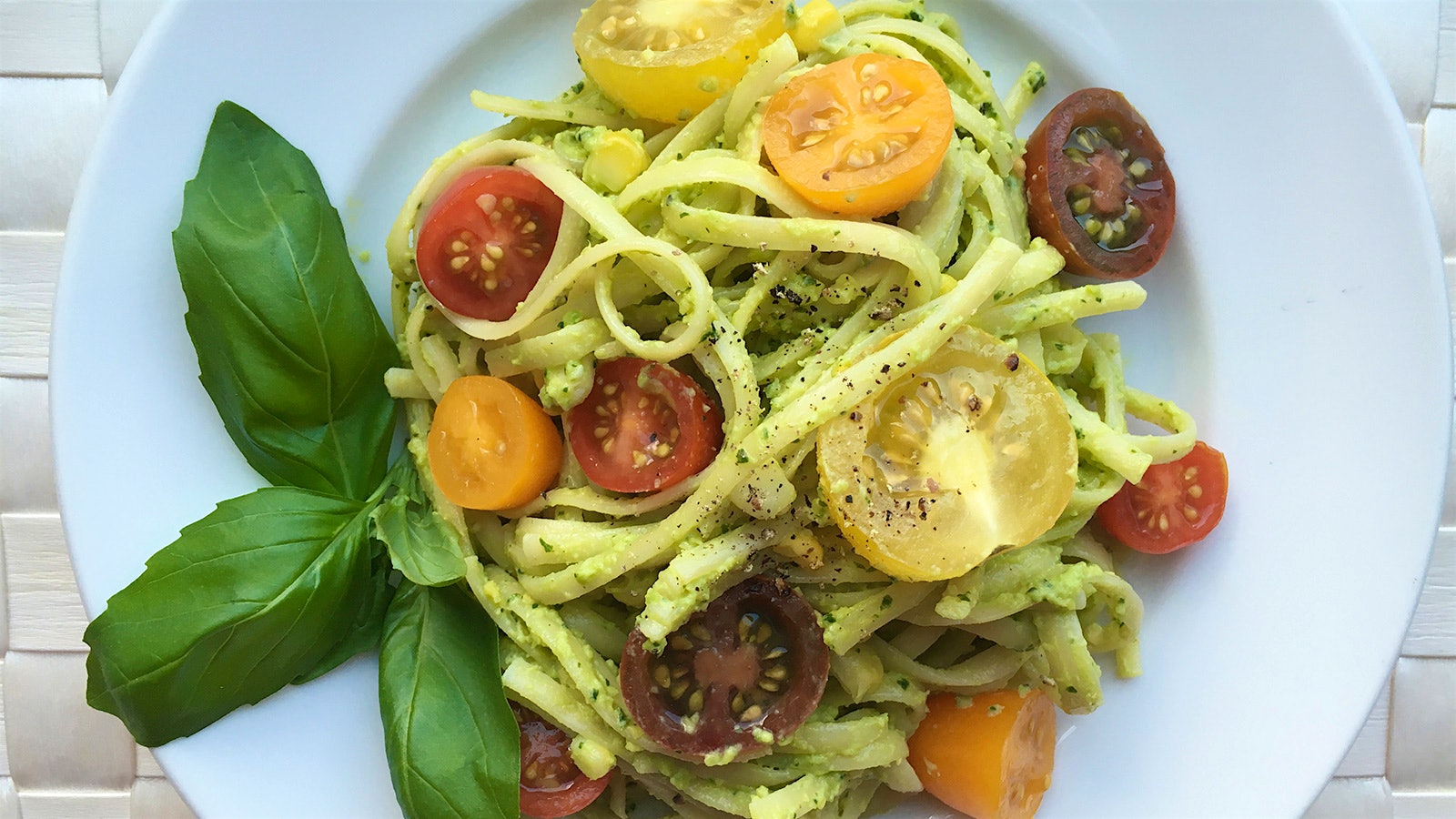 Bowl of pasta with green avocado basil sauce mixed in with colorful, halved baby tomatoes.